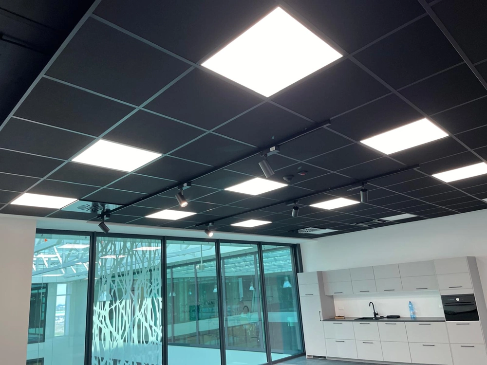 Panele LED w czarnym suficie kasetonowym nad kuchnią biurową z białą zabudową i przeszkloną ścianą