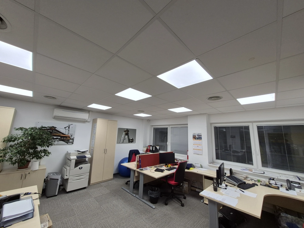 Biuro z sufitowymi panelami LED, dwoma stanowiskami pracy, drukarką i rośliną doniczkową – oświetlenie biurowe