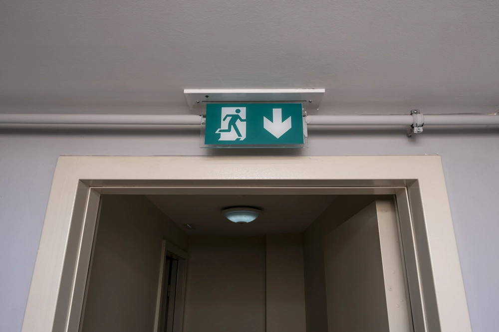 Zielona oprawa oświetleniowa ewakuacyjna z piktogramem biegnącej postaci i strzałką zamontowana nad drzwiami