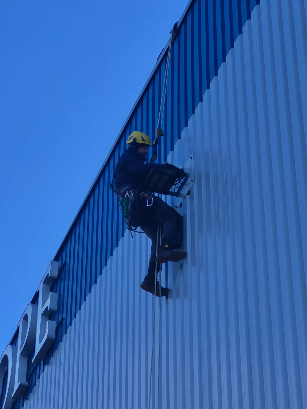 Technik na linach wykonuje montaż oświetlenia na elewacji hali przemysłowej