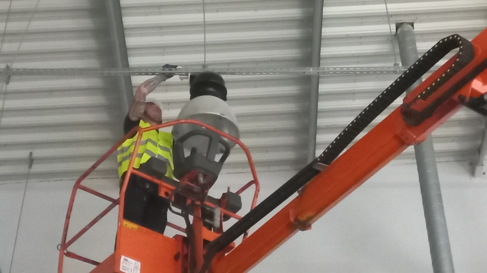 Montaż oświetlenia – technik na podnośniku instaluje dużą lampę przemysłową w hali magazynowej