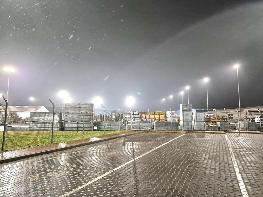 Parking magazynowy z metalowymi rusztowaniami za ogrodzeniem oświetlony naświetlaczami na wysokich słupach, Naświetlacze led